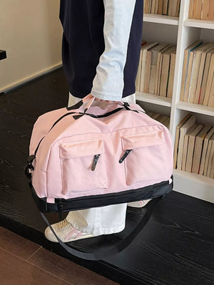 Pink duffel bag with front zip pockets and black trim, held by a person wearing white pants and sneakers.