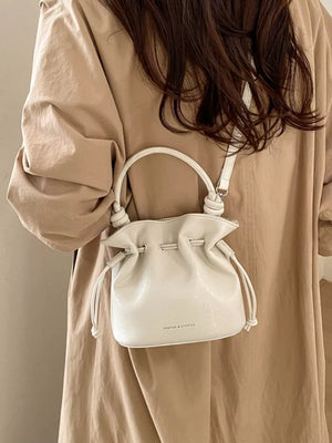 White pu leather cinch-top crossbody bag with knotted handles and drawstring closure.