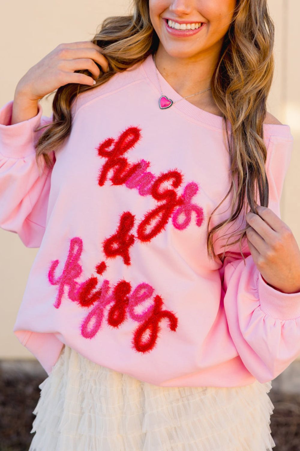 Off-shoulder blush pink sweatshirt with fuzzy red and pink "hugs & kisses" graphic and puff sleeves.