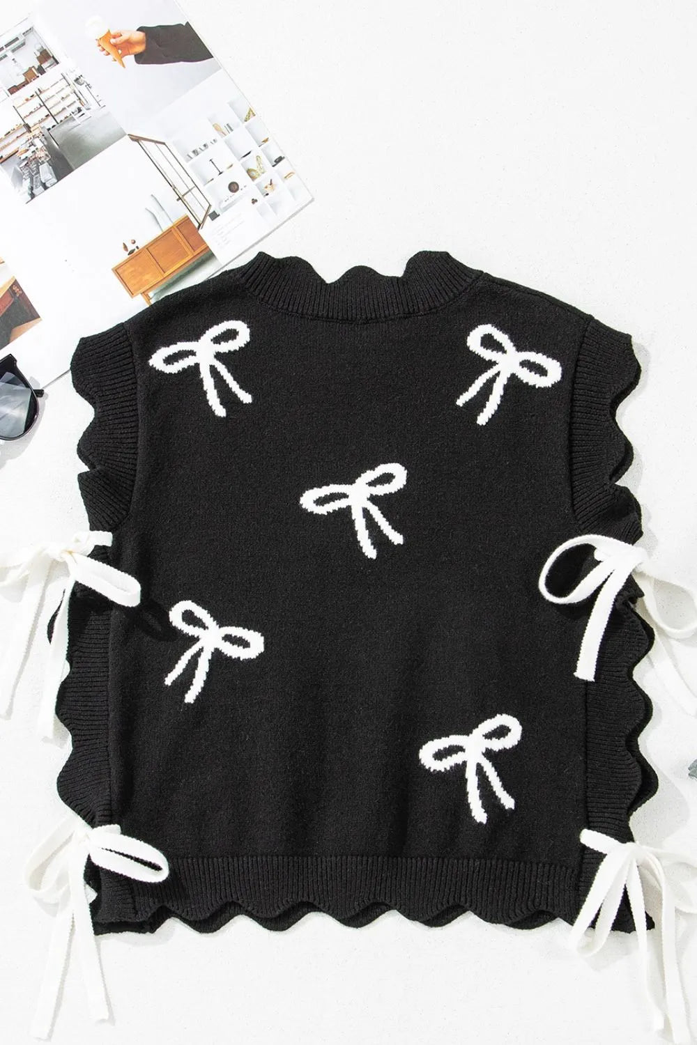 Black knit vest with white bow print, scalloped hem, and tie details on the sides.