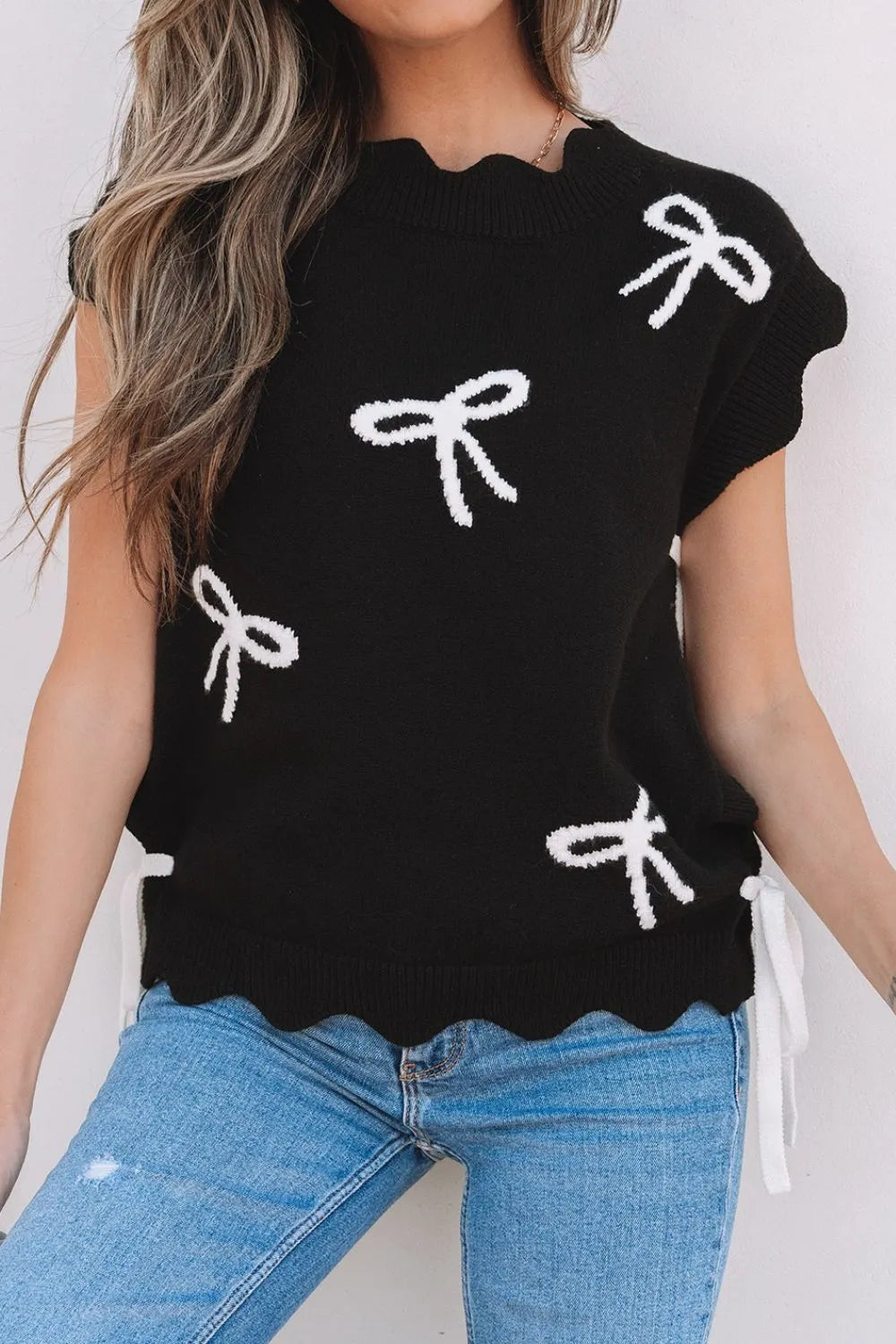 Black knit vest with white bow print, scalloped hem, and tie details.
