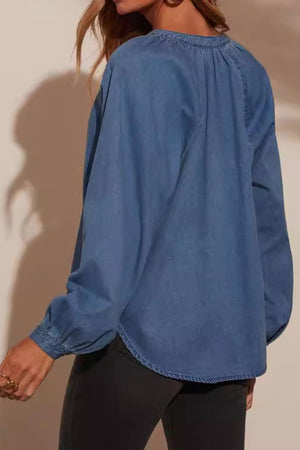 Denim blouson blouse in medium blue with gathered neckline and button cuff sleeves.
