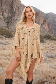 Tan knit poncho with geometric cutouts and fringe, featuring a tie-neck and collar.