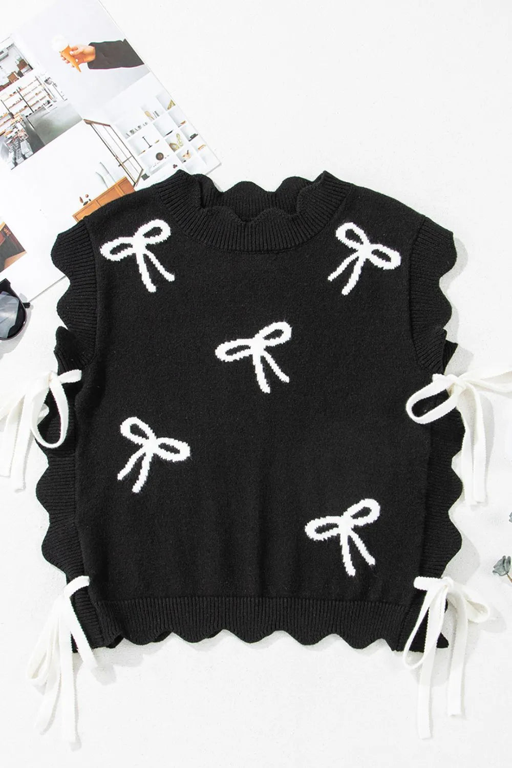 Black knit vest with white bow print, scalloped hem, and tie details.