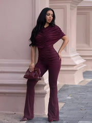Burgundy ruched romper with cap sleeves and flared legs, styled with a clutch bag.