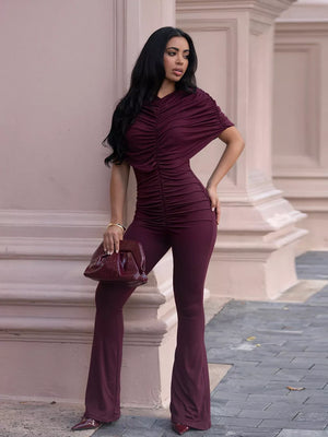 Burgundy ruched romper with cap sleeves and flared legs, styled with a clutch bag.