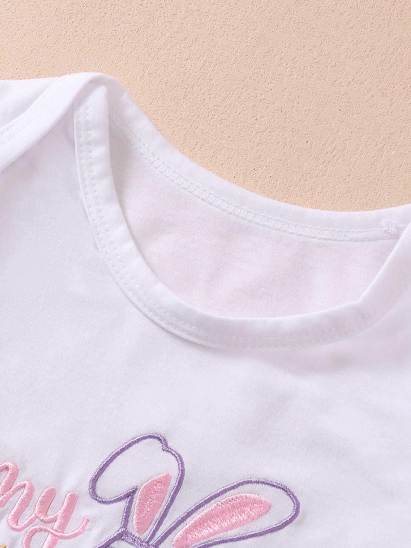 White cotton baby bodysuit with pink and purple bunny embroidery and text.