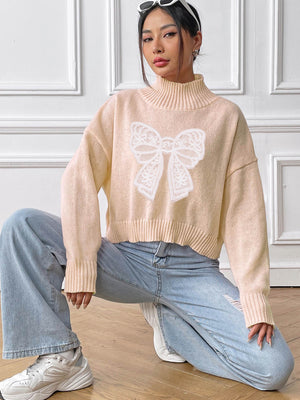 Blush pink turtleneck knit sweater with white lace bow appliqué detail.