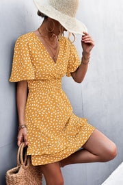 Mustard smocked waist mini dress with white speckle print, V-neck, flutter sleeves, and ruffled hem.