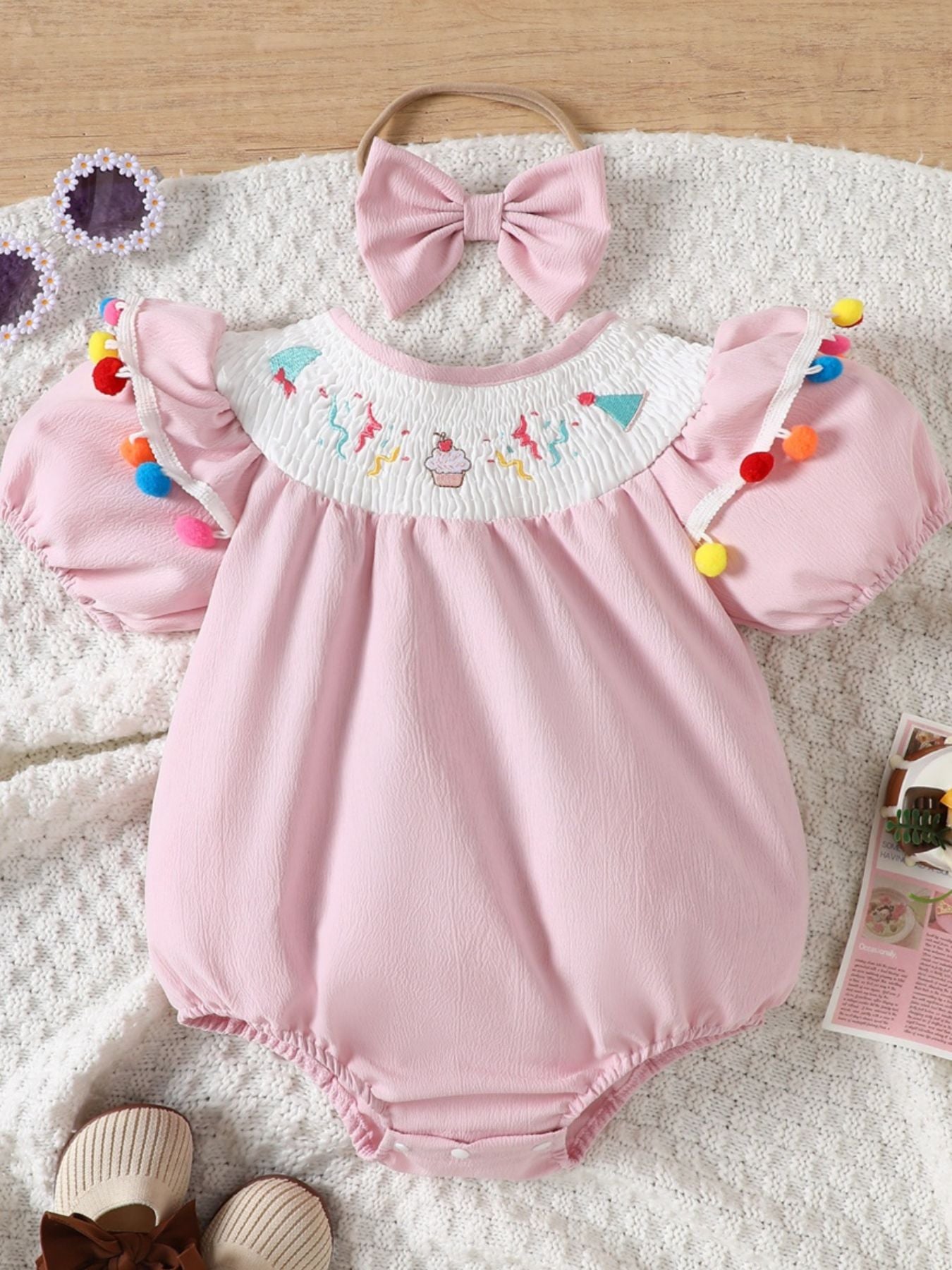 Blush pink cotton baby romper with smocked birthday motif neckline and pom-pom trim.