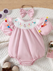 Blush pink cotton baby romper with smocked birthday motif neckline and pom-pom trim.