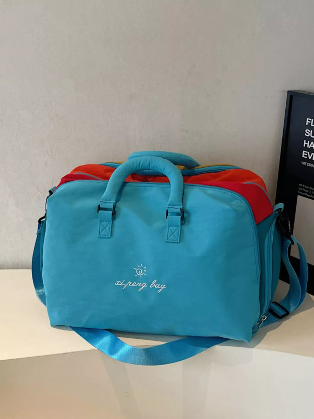 Blue duffel bag with embroidered text and colorful accents, featuring top handles and a shoulder strap.