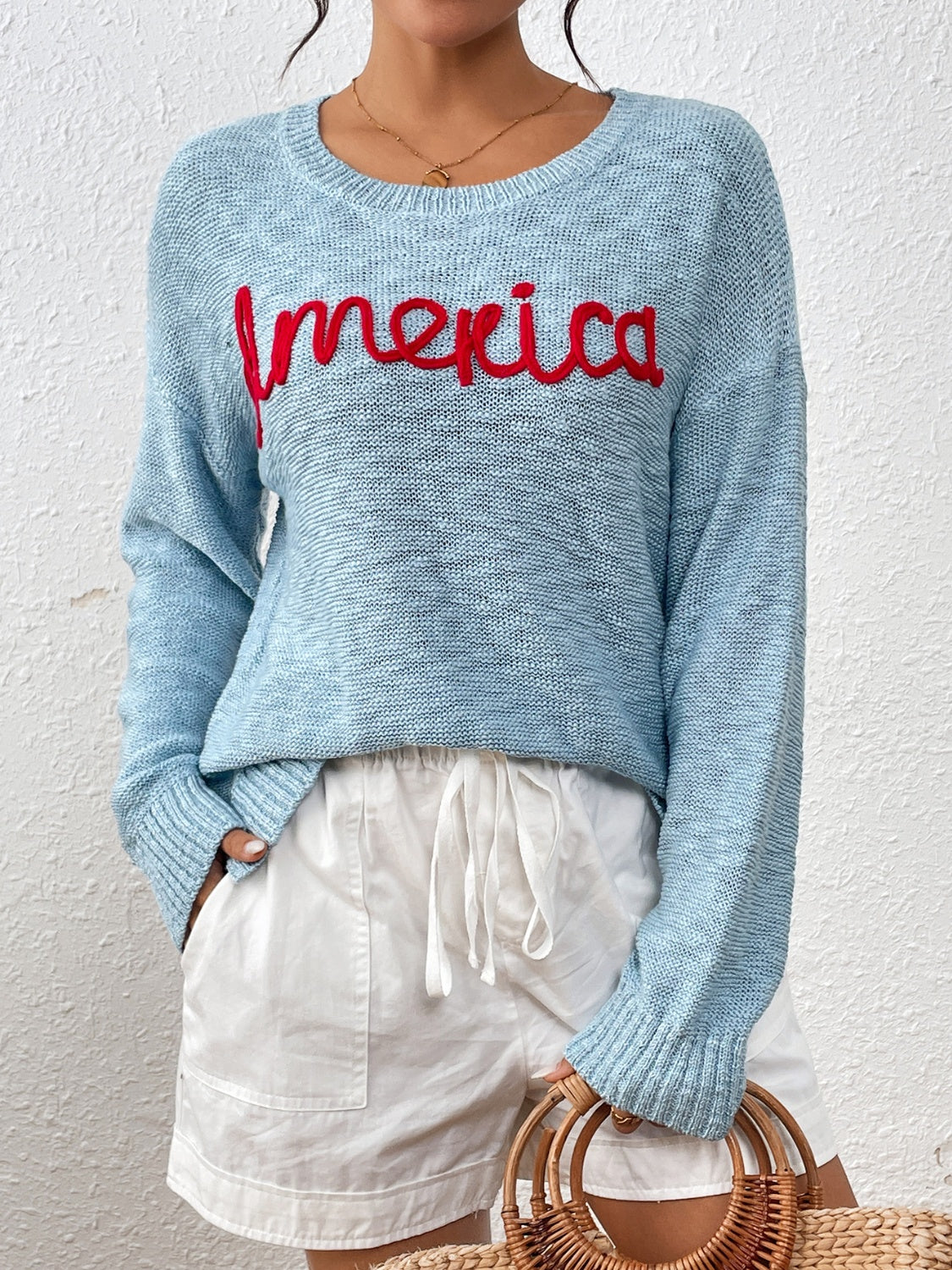 Pastel blue knit sweater with red embroidered "America" text, crew neck, long sleeves.