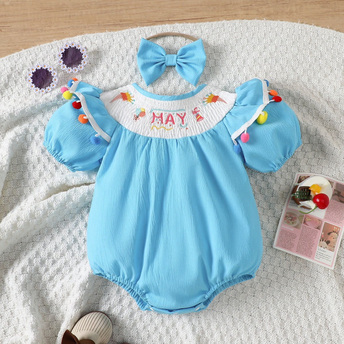 Pastel blue cotton baby romper with embroidered "MAY" and pom-pom trim.