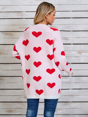Pink acrylic knit cardigan with allover red heart intarsia pattern and white hem.
