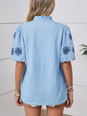 Light blue textured blouse with embroidered floral sleeves and ruffled mock neck.