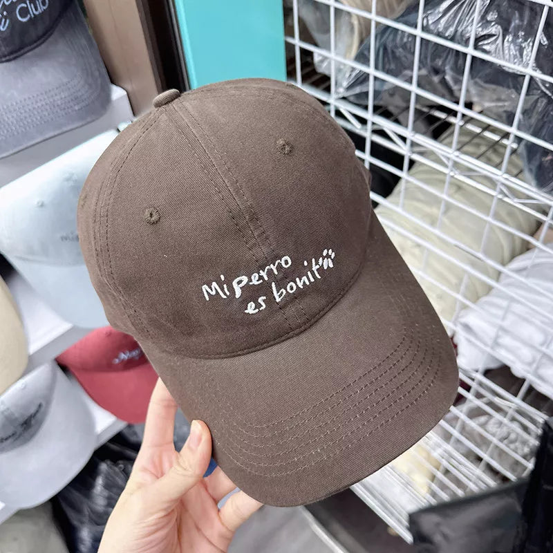 Brown baseball cap with white embroidered "Mi perro es boniti" text and paw prints.