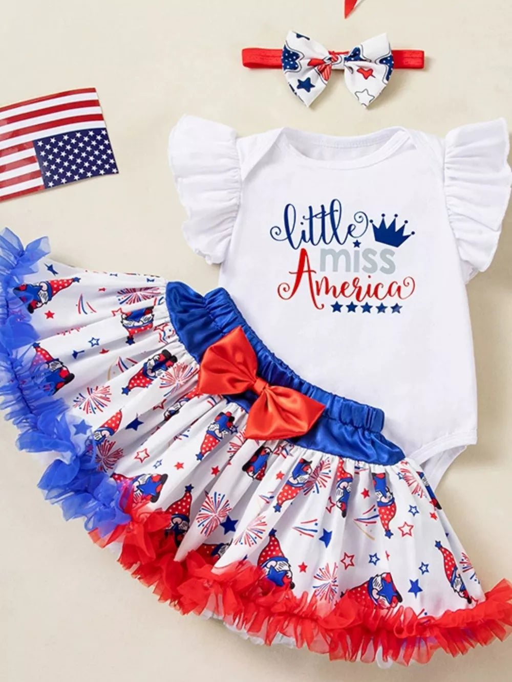 White baby bodysuit with "little miss America" graphic, tiered gnome print skirt with red bow, and patriotic headband.