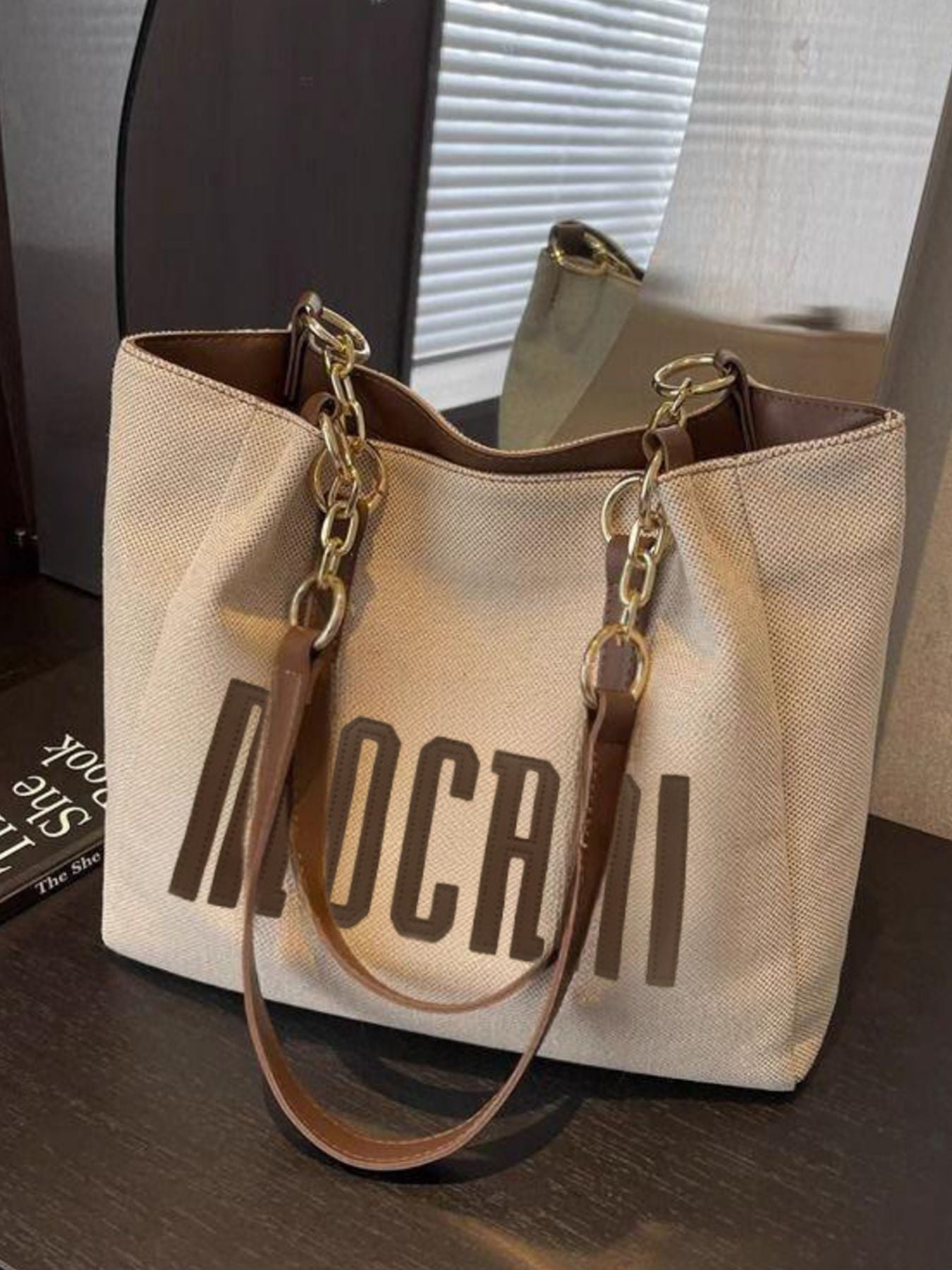 Textured canvas shoulder bag in beige with brown lettering and gold chain accents.