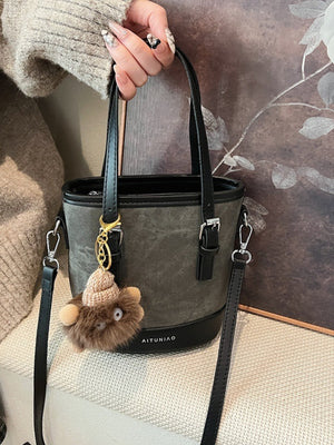 Two-tone bucket crossbody bag in gray with black trim, featuring a fuzzy charm and detachable strap.