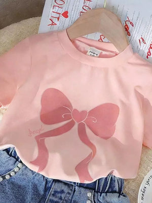 Blush pink tee with bowknot graphic and text, paired with blue denim shorts with pearl details.