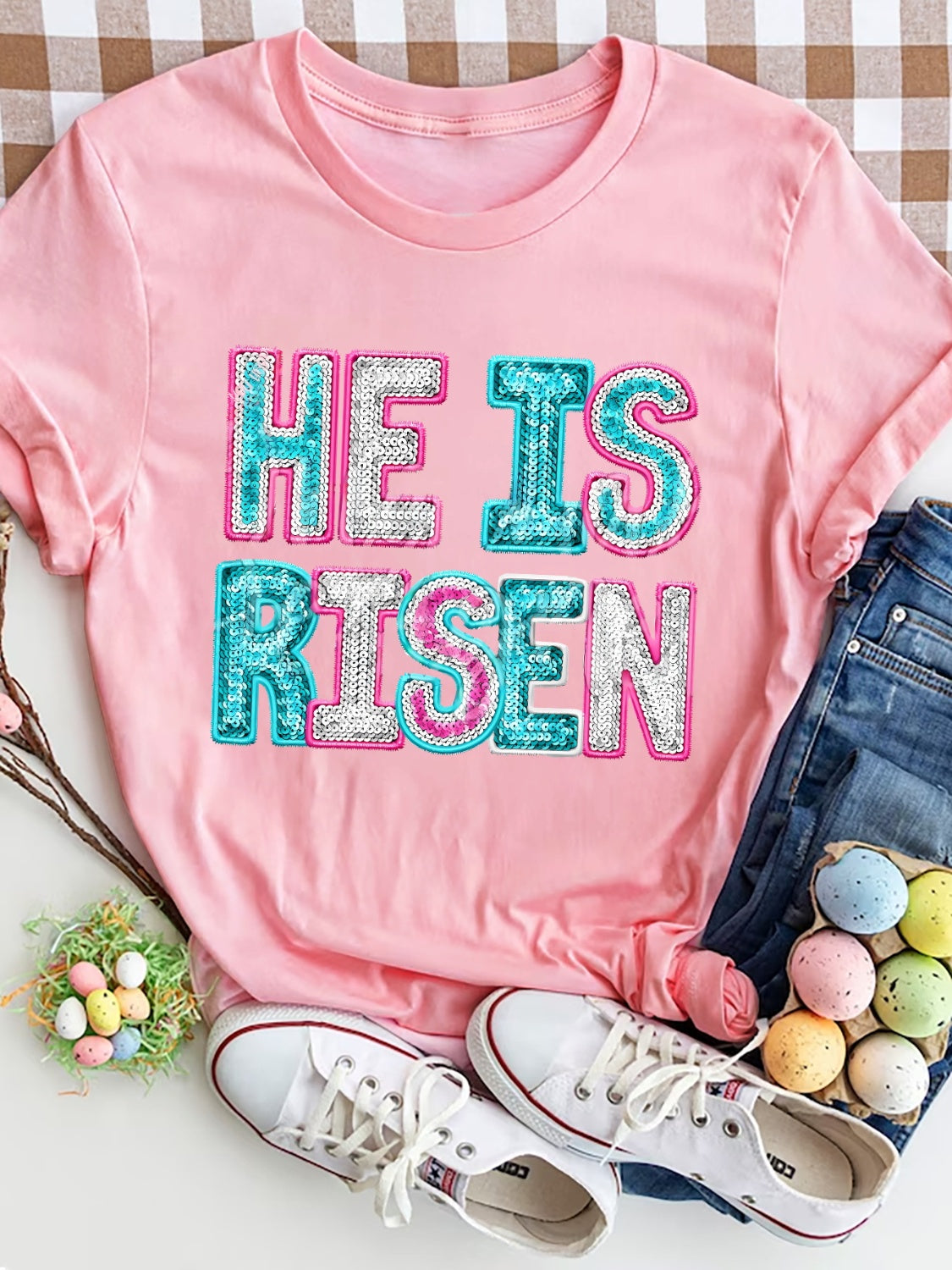 Pink crewneck t-shirt with sequin "He Is Risen" graphic.