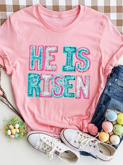 Pink crewneck t-shirt with sequin "He Is Risen" graphic.
