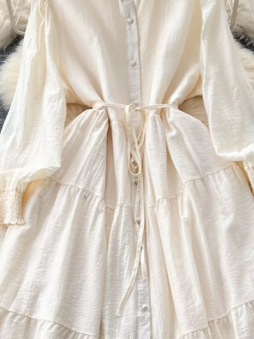 Off-white tiered long sleeve button-down dress with tie waist and textured fabric.