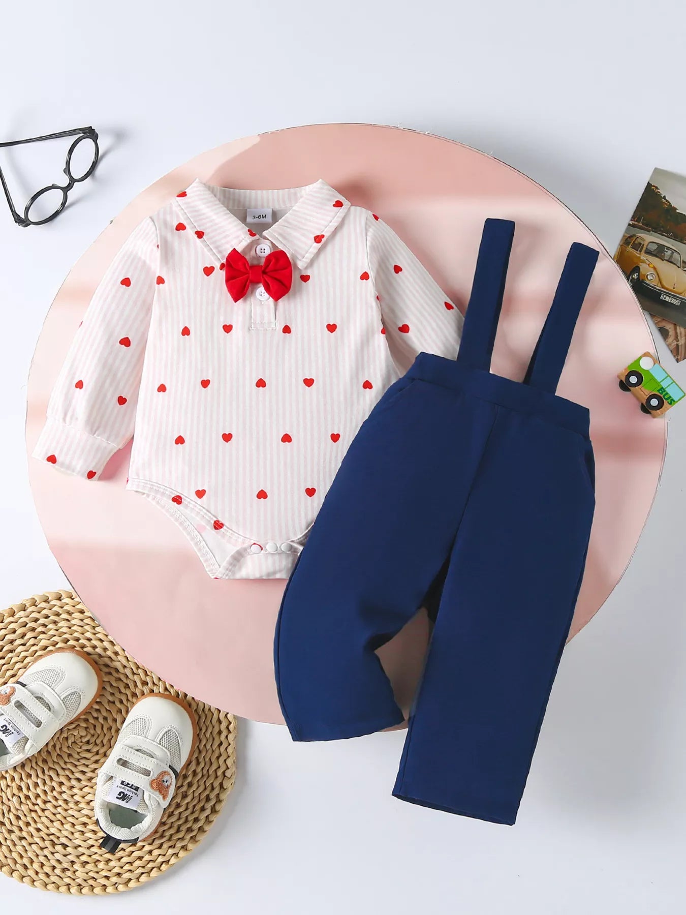 White striped baby bodysuit with red hearts and red bow tie, paired with navy suspender pants.