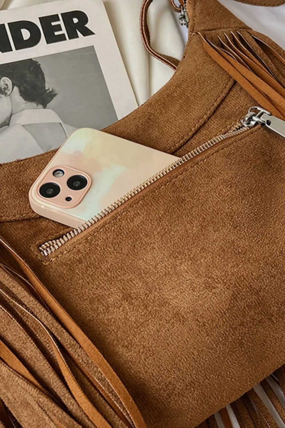 Brown suede crescent crossbody bag with fringe detail and zip pocket holding a phone.