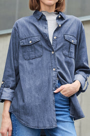 Relaxed denim shirt in navy with patch pockets and buttoned cuffs, worn over a grey top.