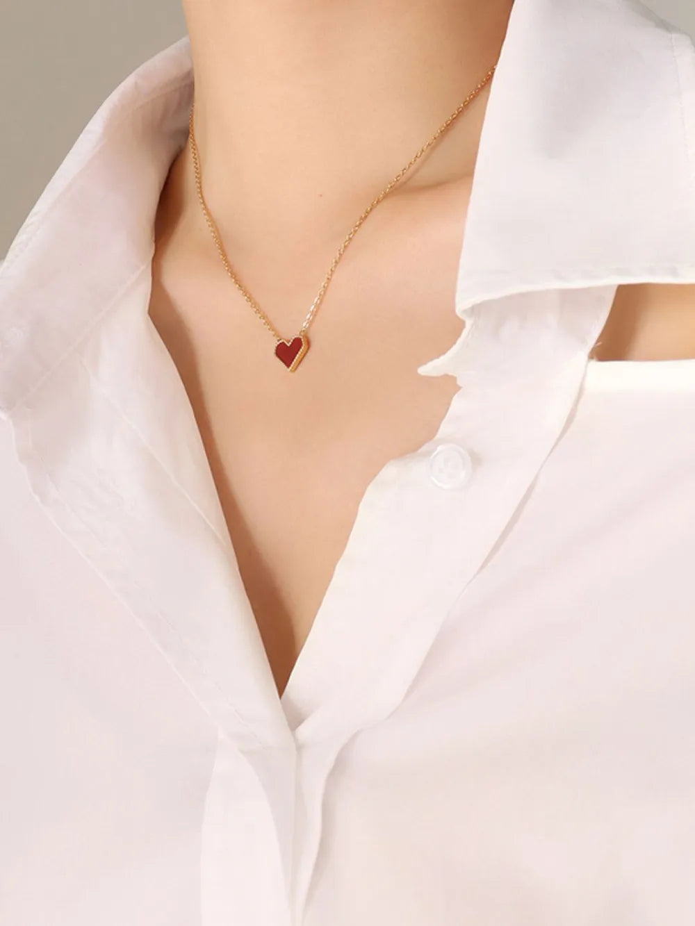 Gold necklace with a small crimson heart pendant on a white collared shirt.