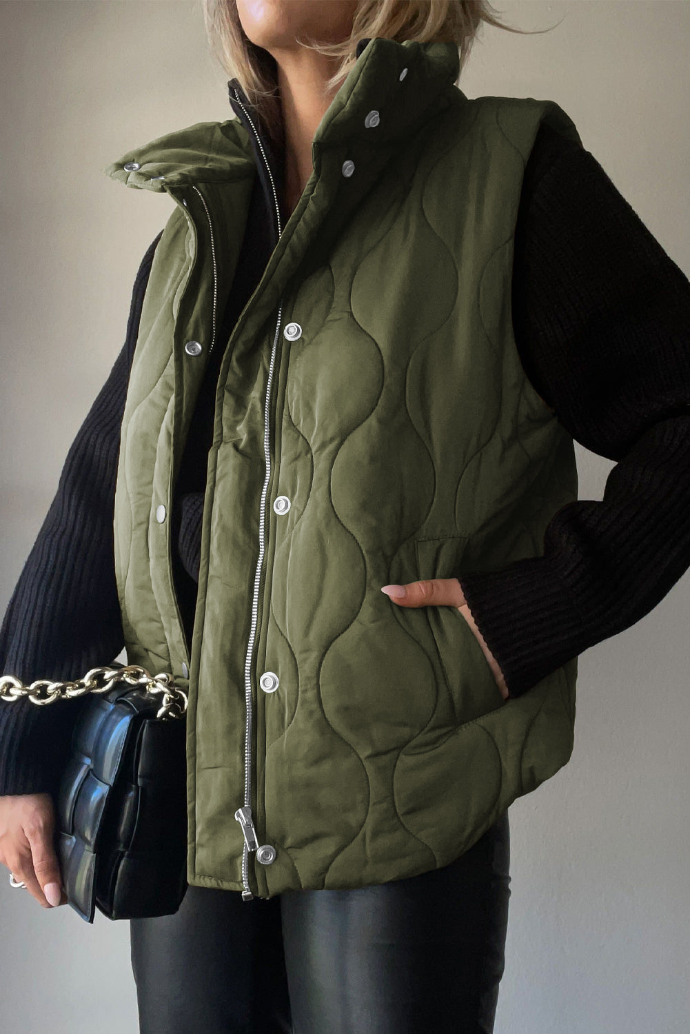 Green quilted stand-collar utility gilet with snap and zip closure, styled with a black sweater and bag.