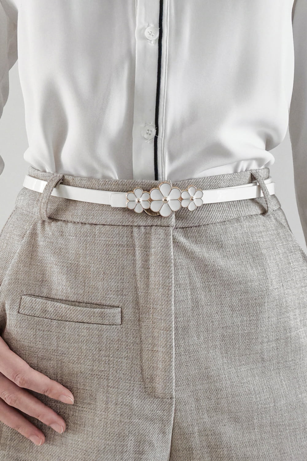 White skinny belt with floral blossom buckle detail, styled with a white shirt and grey trousers.