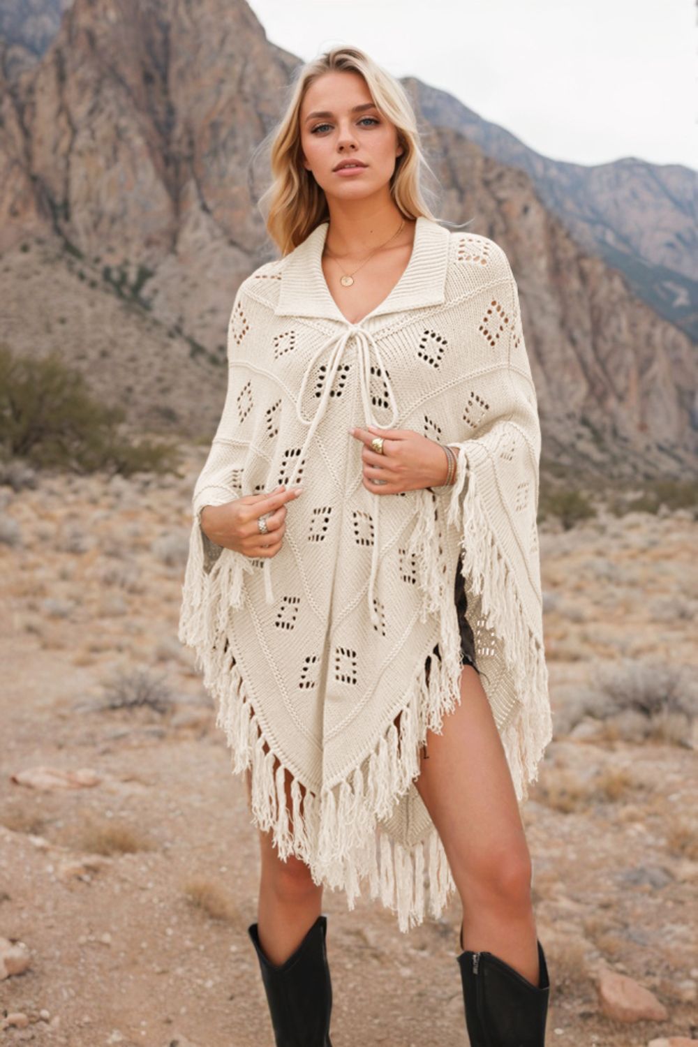 Cream knit poncho with geometric pattern, fringe, and tie neck, styled with black boots.