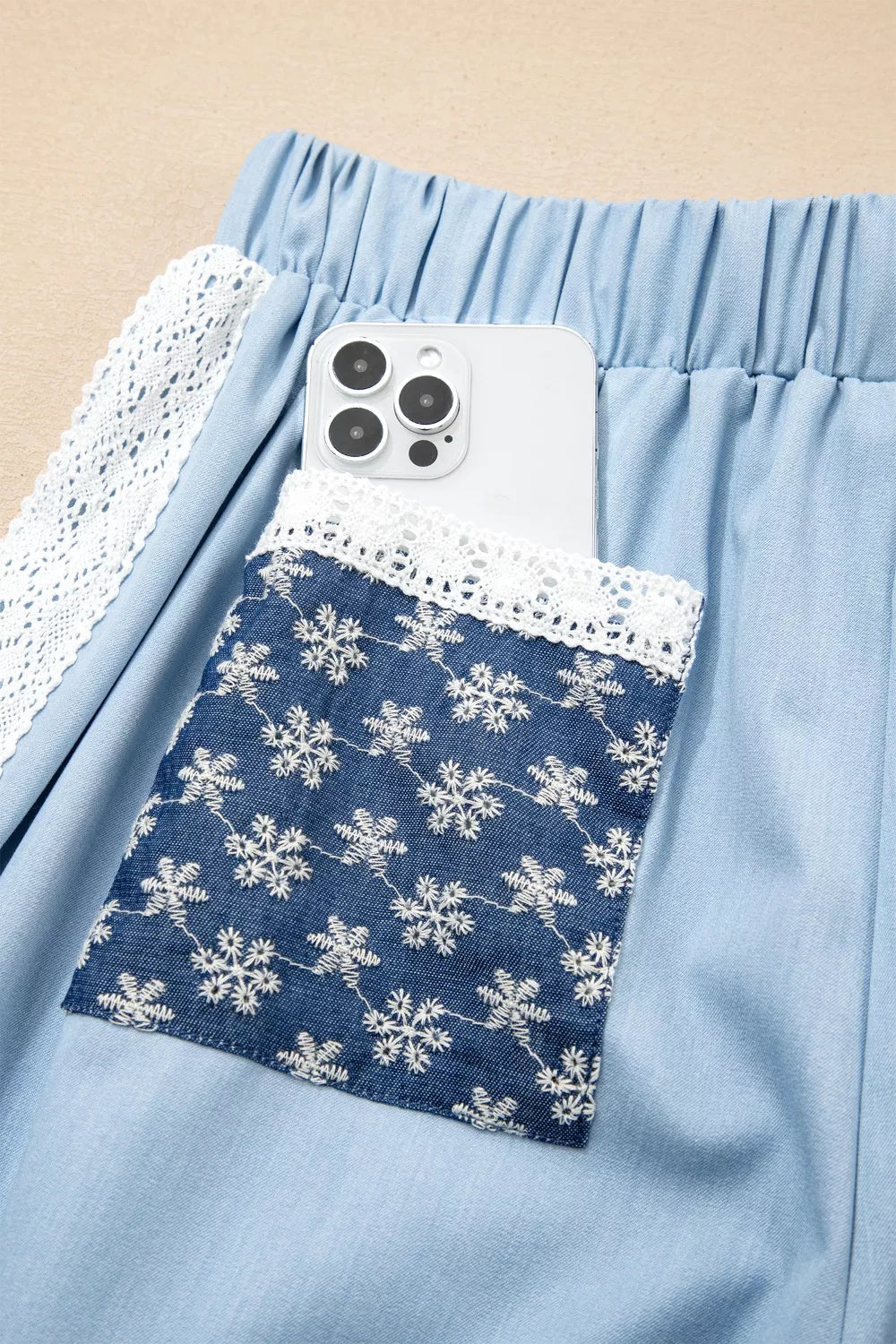 Light blue maternity pants with drawstring waist, lace trim, and embroidered snowflake pocket holding a phone.