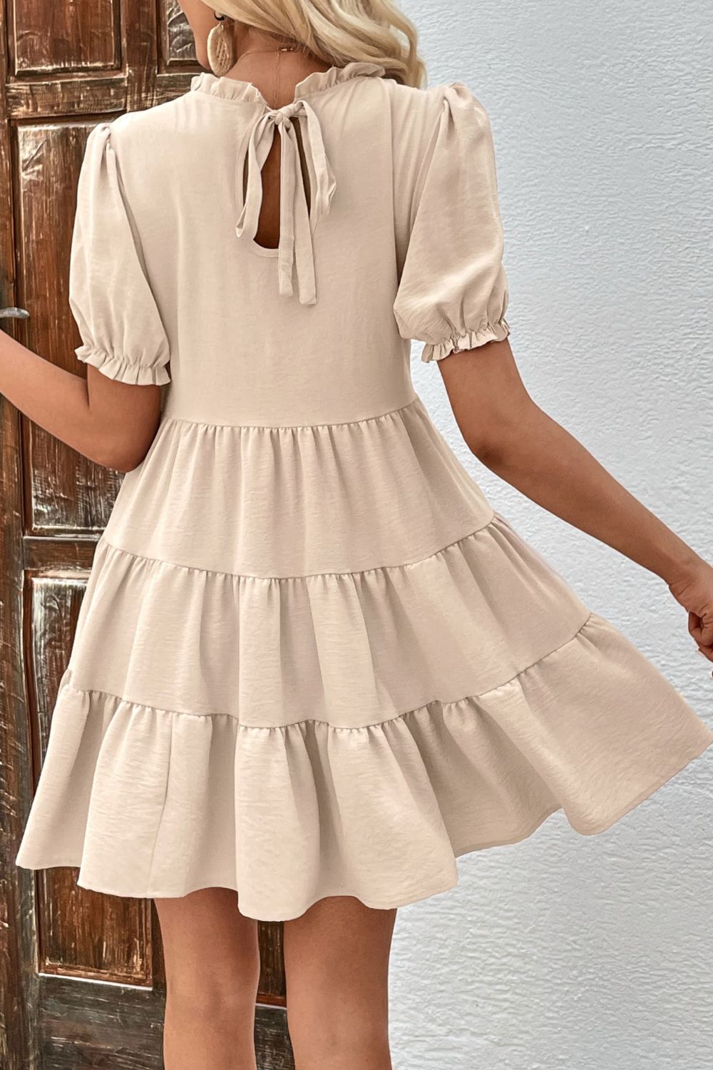 Beige tiered mini dress with ruffled neckline, puff sleeves, and tie back detail.