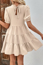Beige tiered mini dress with ruffled neckline, puff sleeves, and tie back detail.