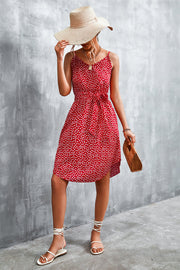 Red daisy print slip dress with tie waist and spaghetti straps, styled with a straw hat and sandals.
