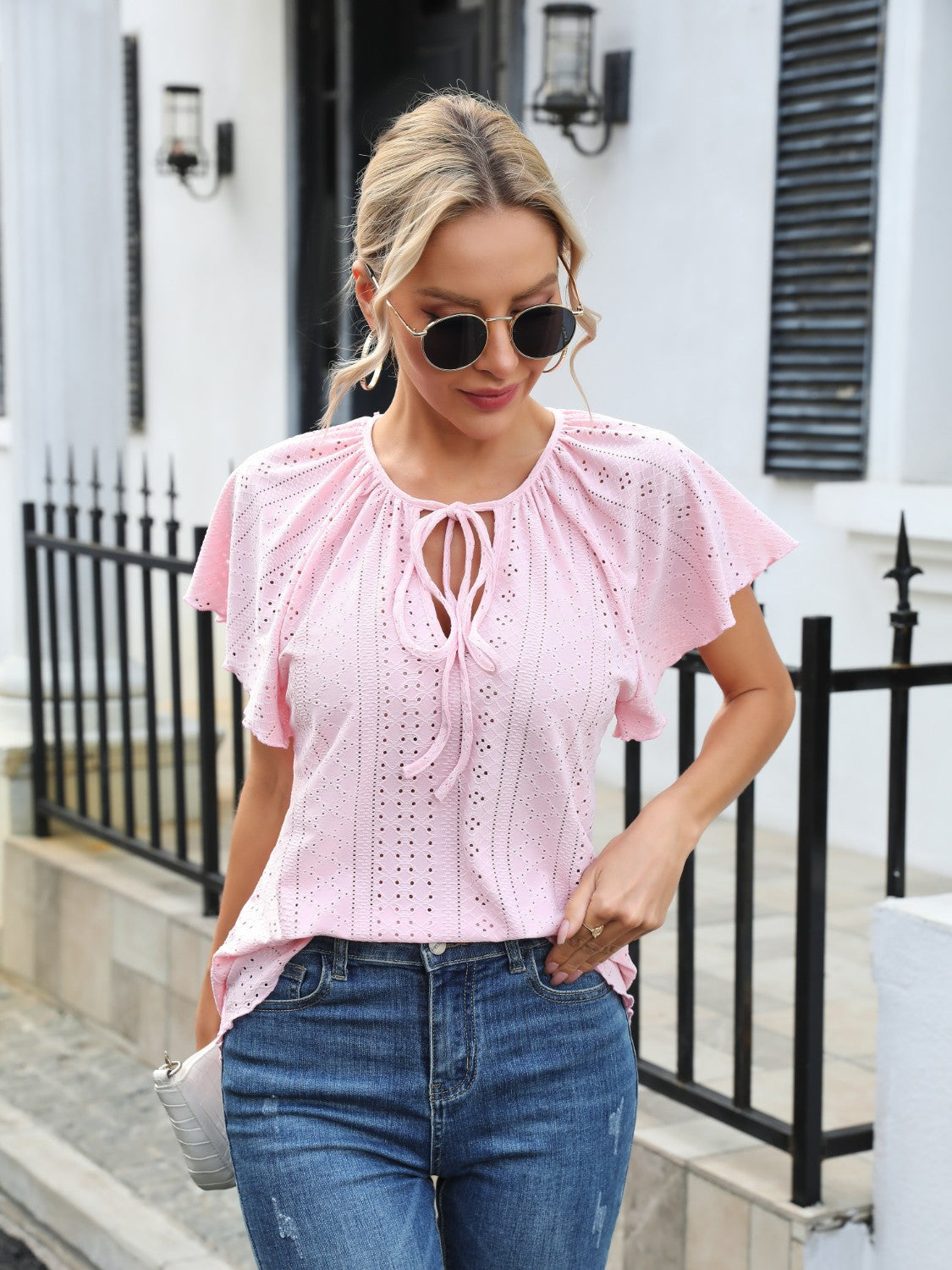 Blush pink perforated tie-neck blouse with flutter sleeves and eyelet details.