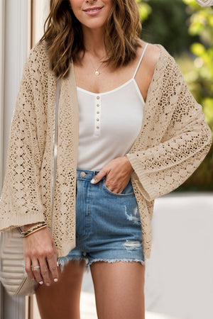 Open-knit crochet cardigan in Dust Storm acrylic with wide sleeves, worn over a white cami and denim shorts.