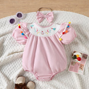 Blush pink baby romper with smocked birthday motif neckline, puff sleeves, and colorful pom-pom trim.
