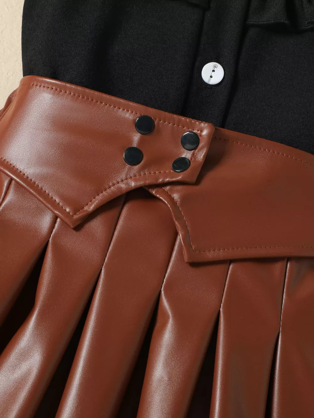 Black baby top with white button detail and brown faux leather pleated skirt with snap closures.