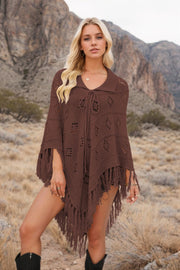 Brown knit poncho with geometric pattern, fringe, and tie neck, styled over shorts.