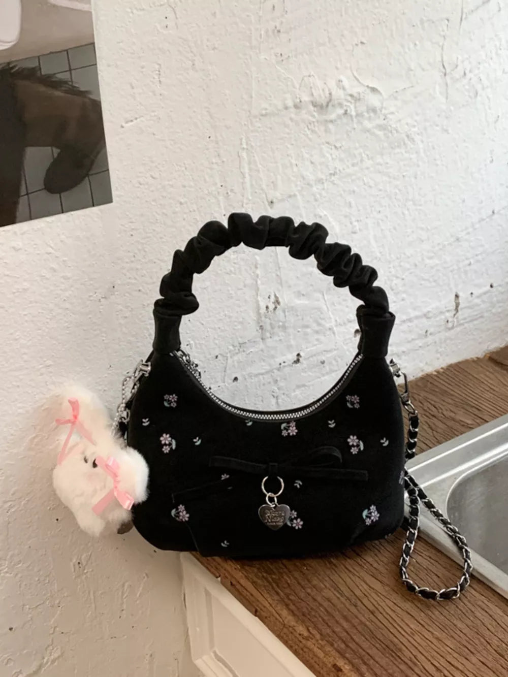 Black ruched mini crossbody bag with floral embroidery and bow charm.