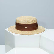 Refined straw boater hat in beige with a brown ribbon detail and small metal emblem.