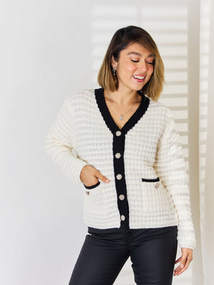 White textured knit cardigan with black contrast trim, buttons, and pocket details.