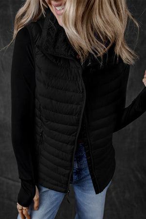 Black quilted puffer vest with soft fleece collar and zip front, worn over a long sleeve top.
