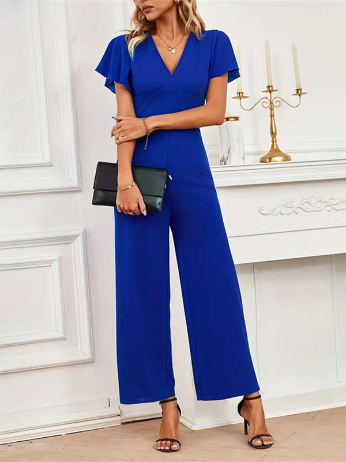 Royal blue flutter sleeve wide-leg jumpsuit with V-neck and clutch bag.