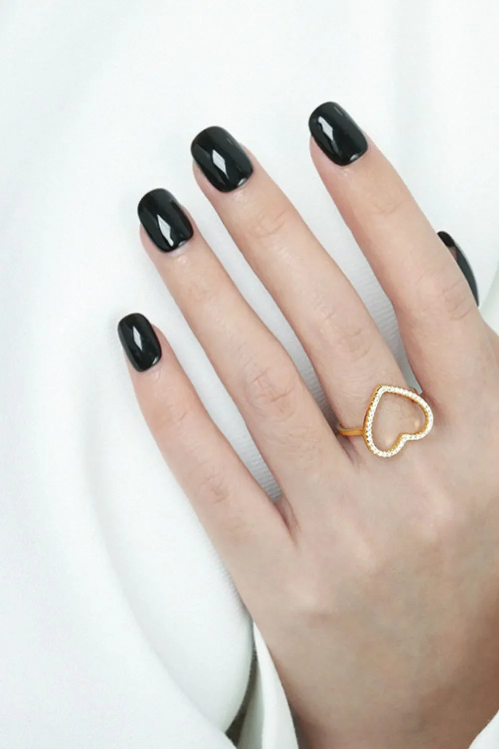 Gold pavé heart outline ring with clear stones on a hand with black nails.
